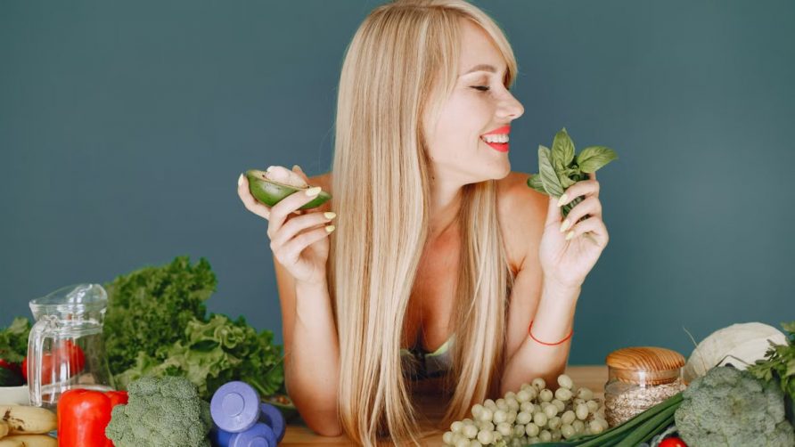 A smiling woman holding avocado and basil, surrounded by fresh vegetables and fruits for healthy eating.