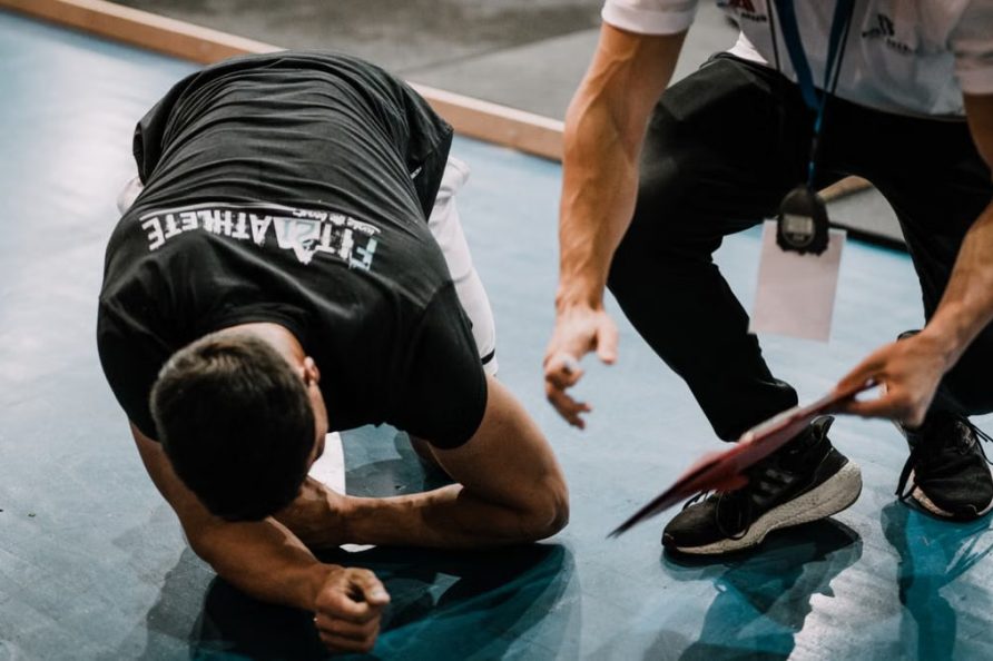 An athlete being assisted by a coach during an indoor training session on a blue floor.